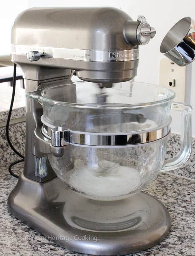 A silver stand mixer with a clear bowl and eggs whites in the bottom.