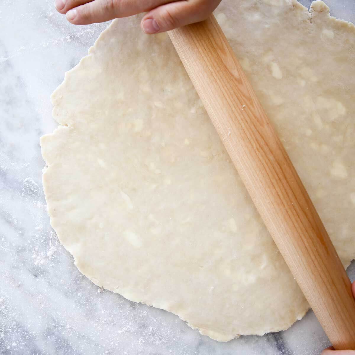 pie dough rolled on marble with hands on rolling pin.