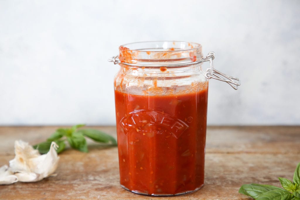 Red wine pasta sauce inside a clear jar on a wood surface with fresh garlic and herbs.