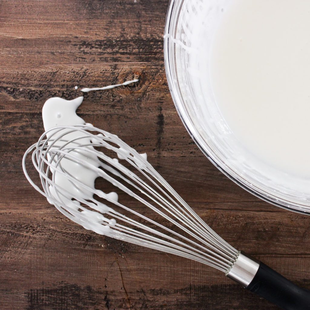 White glaze spilling from a whisk onto a wood board next to a bowl of white glaze.