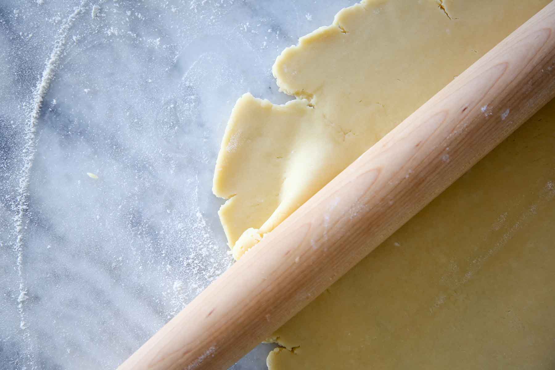 pate sucree dough disk being rolled thin by wooden pin on marble.