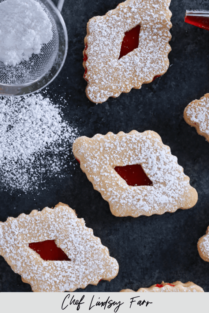 Raspberry linzer cookies laid out on a dark background with powdered sugar sifter nearby.