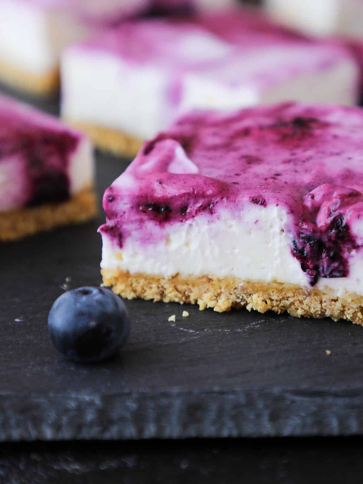 A fresh blueberry next to a square blueberry cheesecake bar.