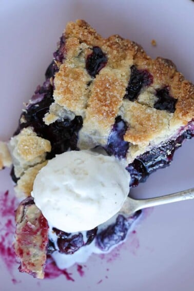 A slice of blueberry pie with a golden lattice top and scoop of vanilla ice cream.