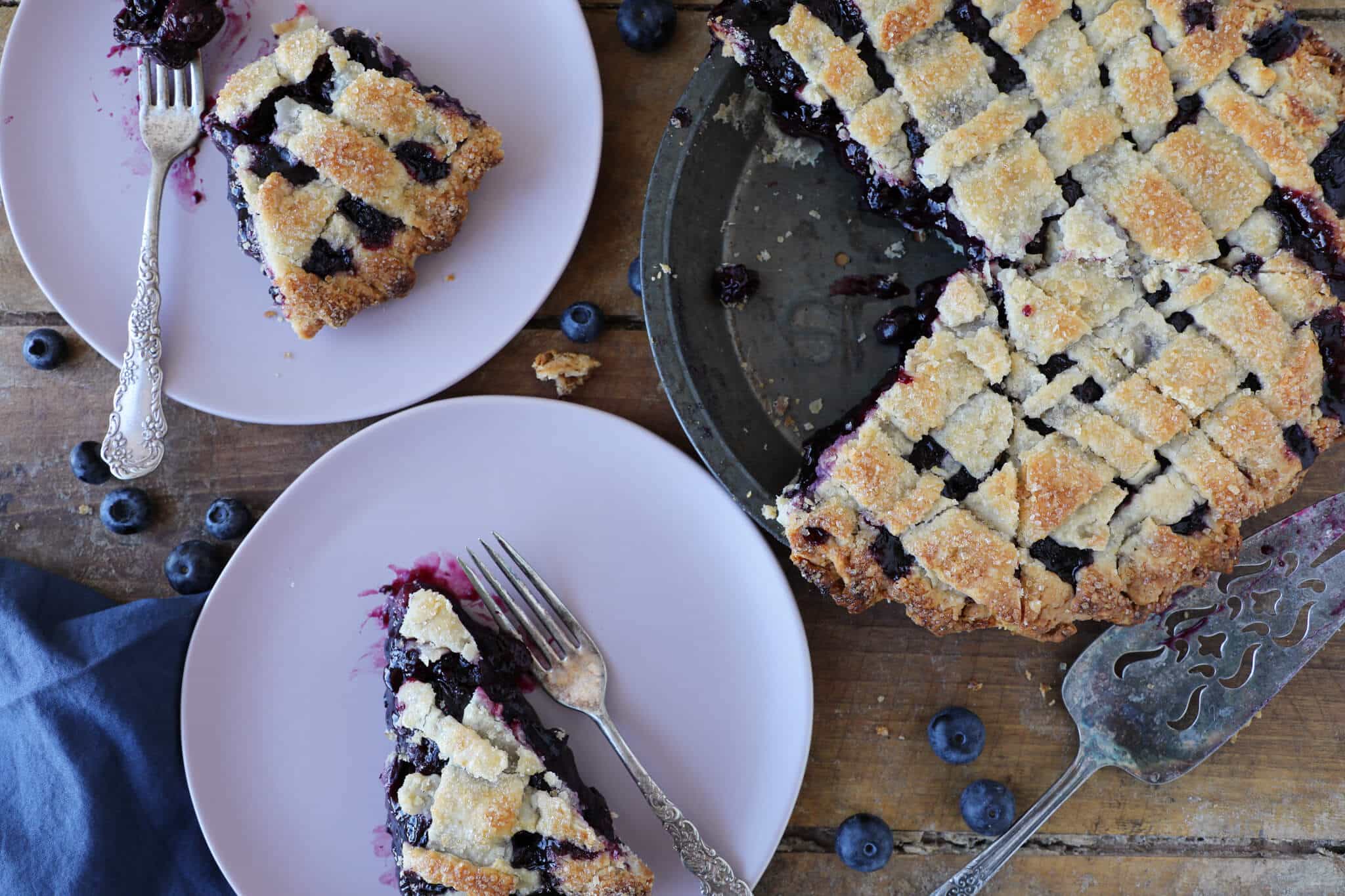 Two servings of blueberry pie next to a whole blueberry pie on a wood surface.