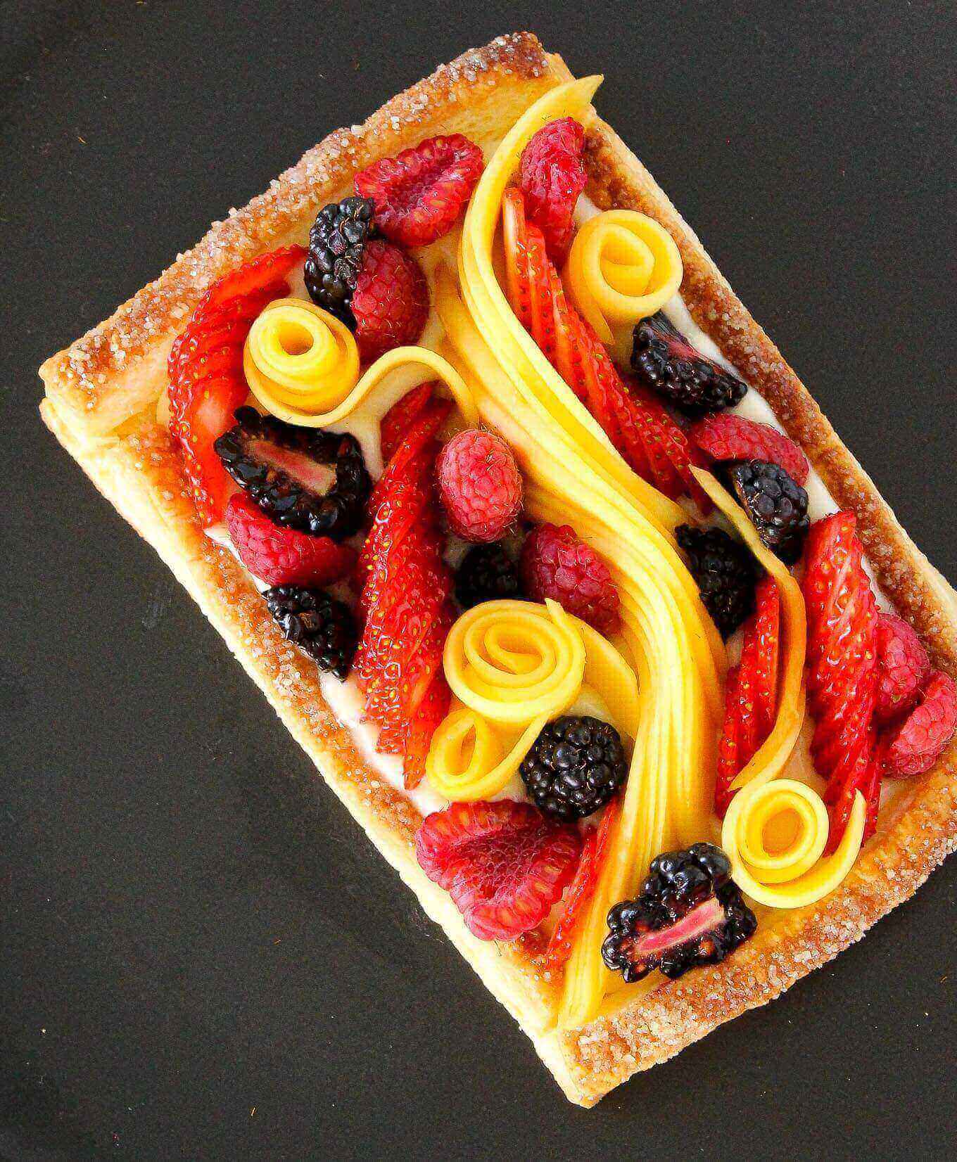 Puff pastry tart with decorative fruit including fresh berries and sliced mango along the center.