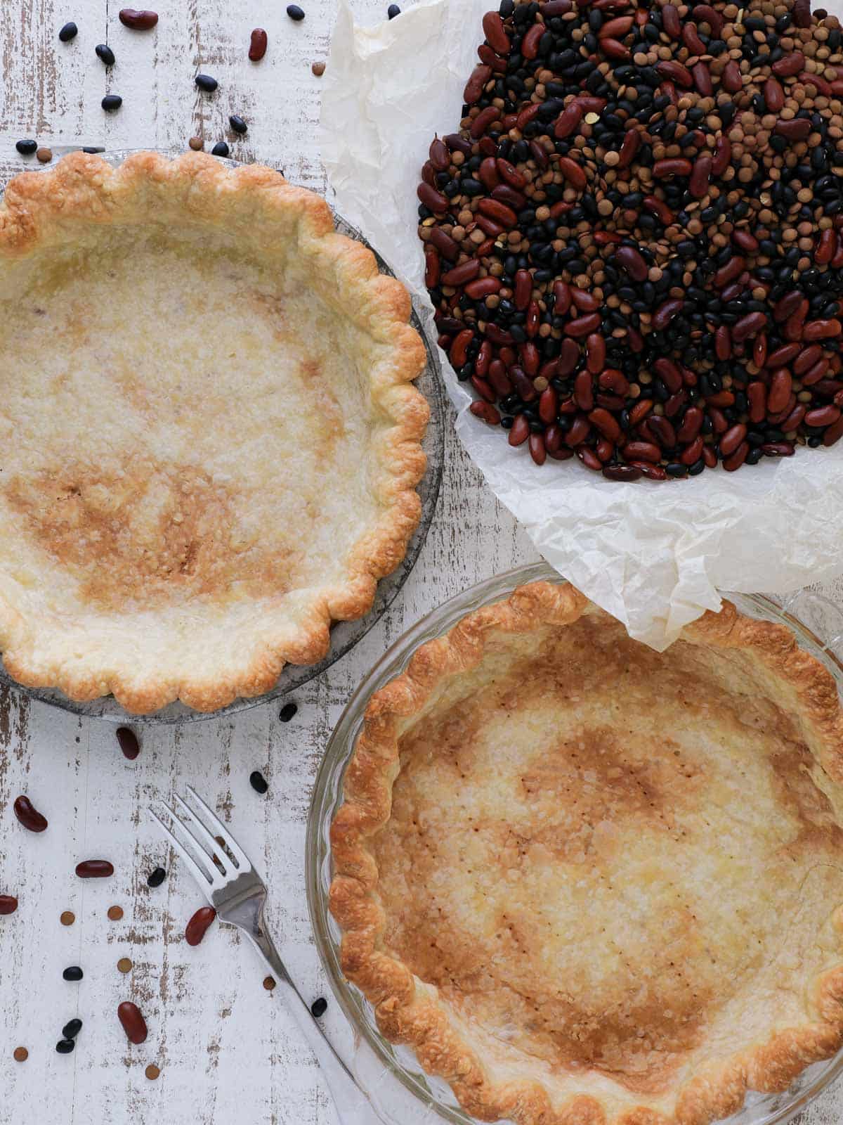 two blind baked pie crusts on white wooden surface with baking beans.