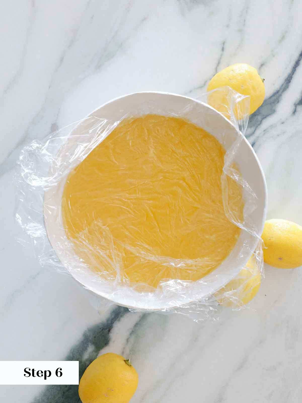 cooling lemon curd with plastic wrap on surface in white bowl.