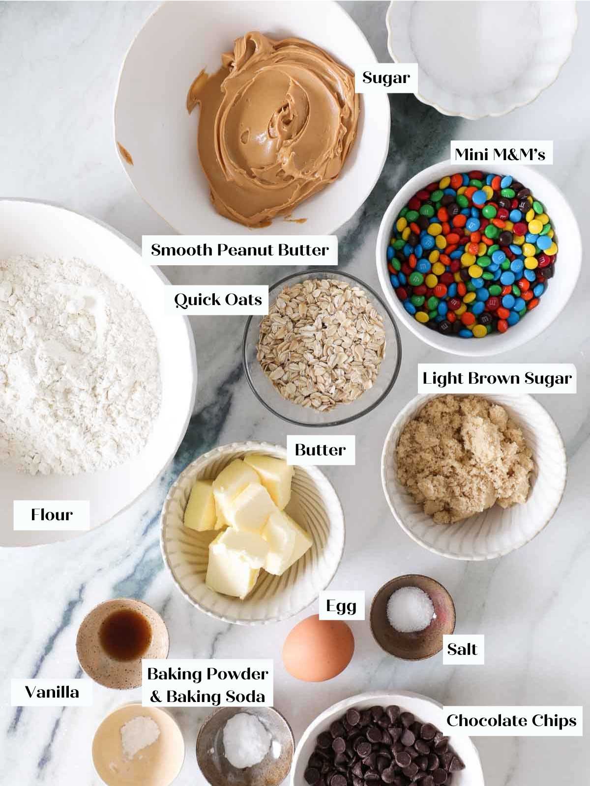 A flat lay of monster cookie ingredients on a white marble counter, including oats, peanut butter, and M&Ms.