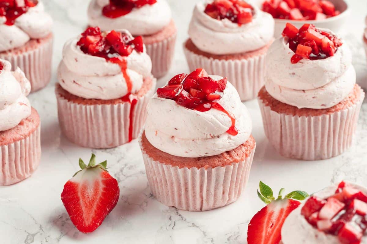 A collection of strawberry cupcakes surrounded by fresh strawberries.