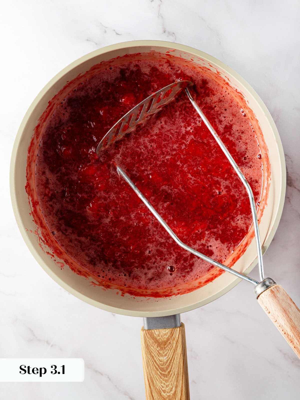 Cooked strawberries being mashed with a fork to create a chunky jam for cupcakes.