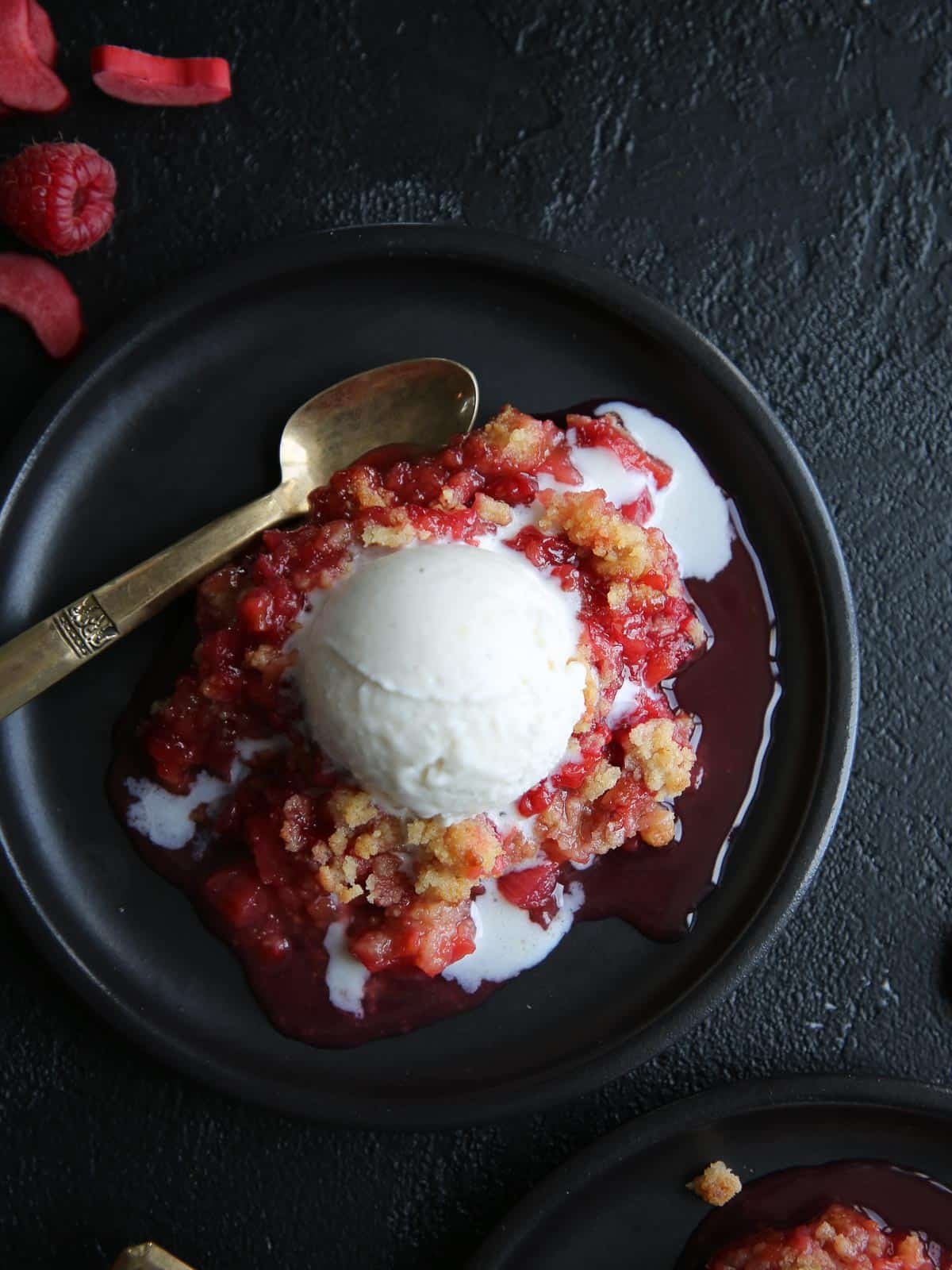 Raspberry rhubarb crisp served on a black plate with a scoop of vanilla ice cream.
