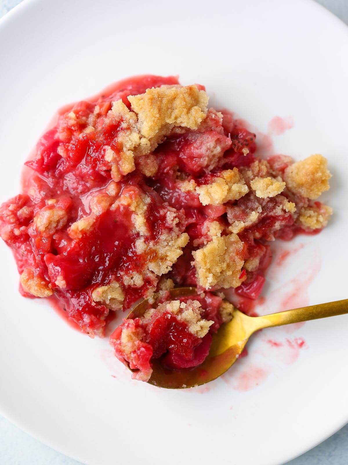 A serving of strawberry rhubarb crumble on a white plate with fruit showing through.