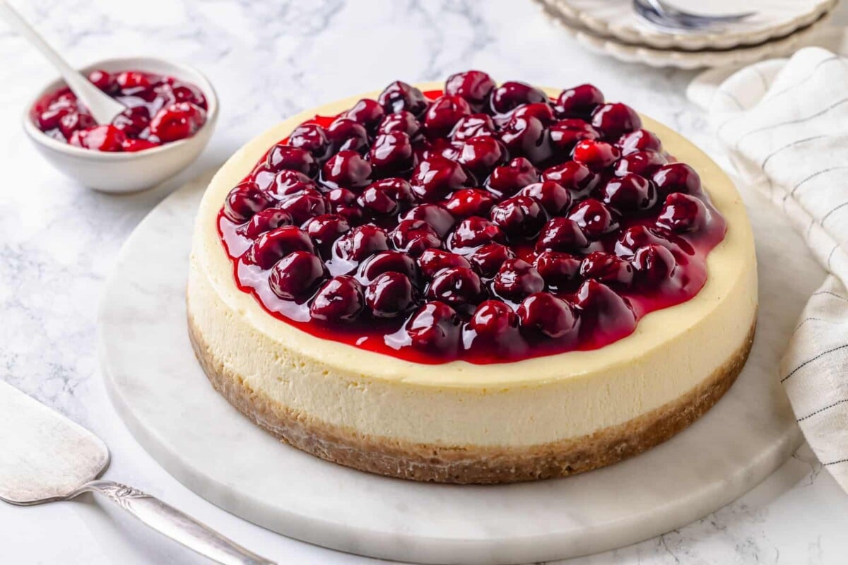 Finished cherry cheesecake resting on a white cake stand placed on a marble surface.