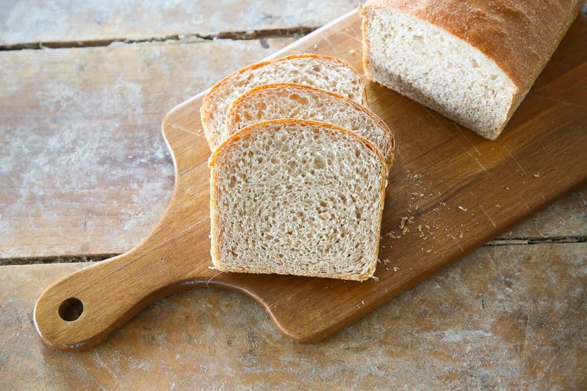 A whole wheat sandwich loaf sliced on a wooden cutting board.