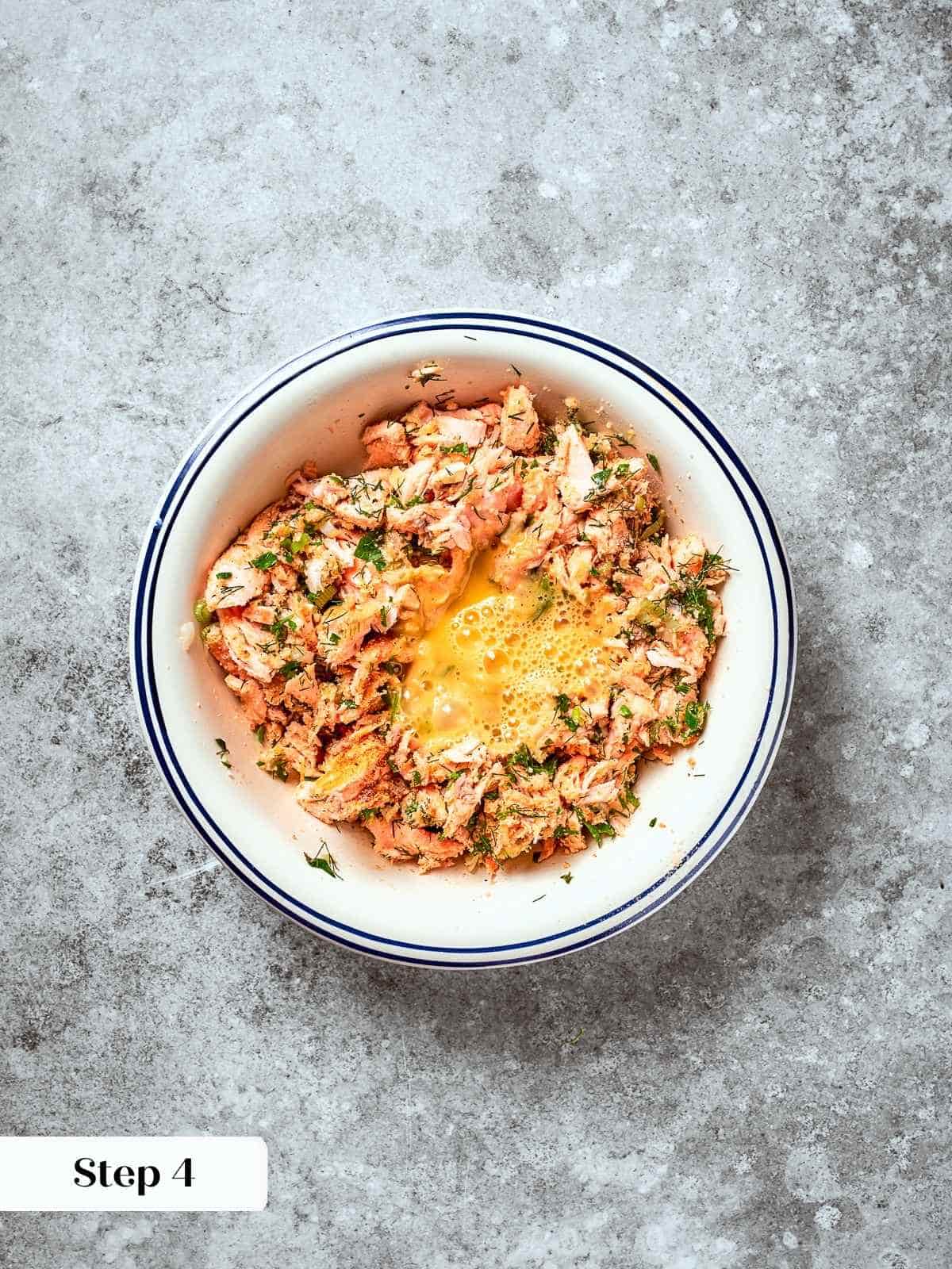 Egg being added to salmon patty mixture in a mixing bowl with herbs and breadcrumbs.