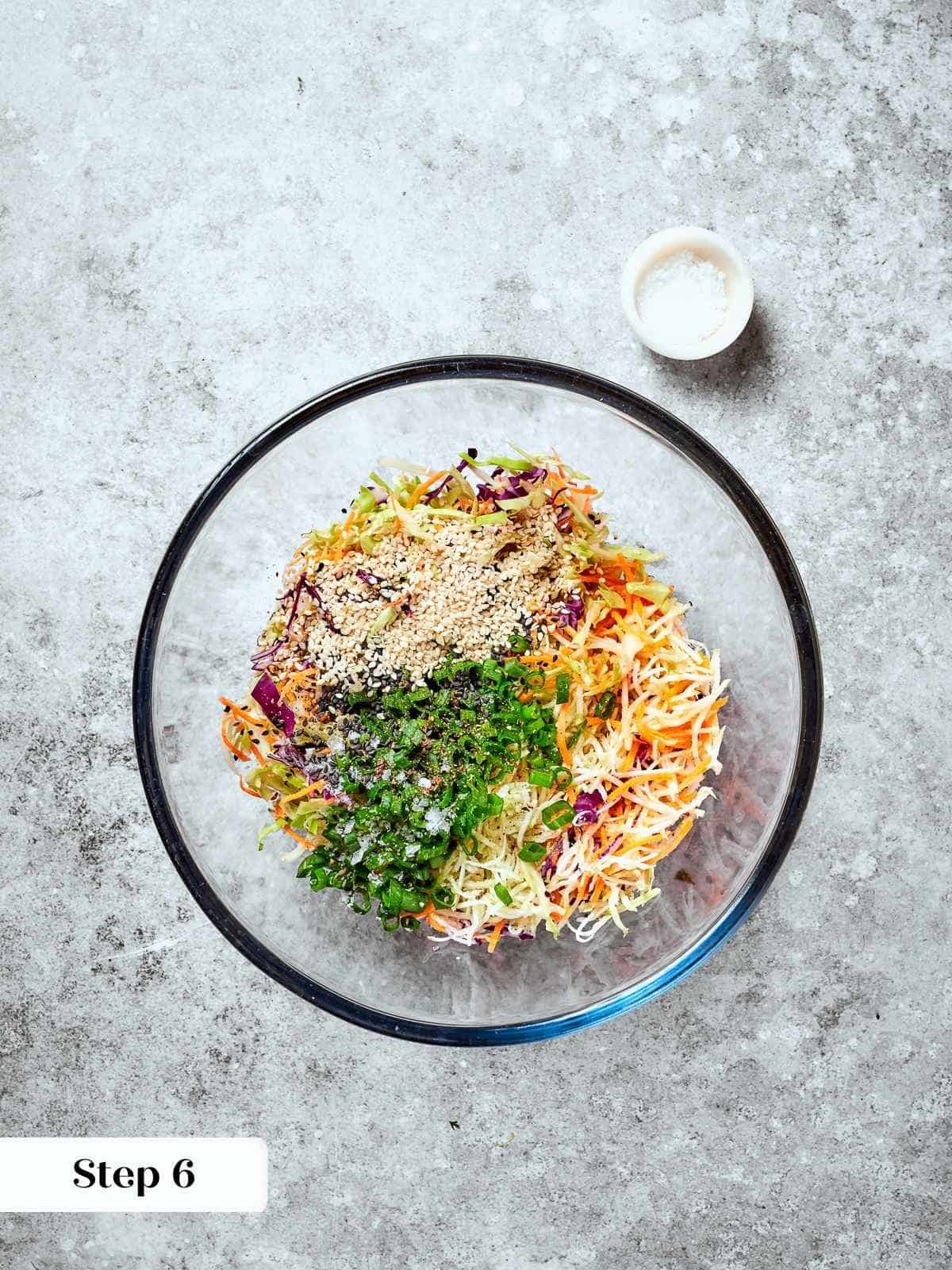 Mixing bowl filled with raw slaw ingredients, including broccoli mix and sesame seeds.