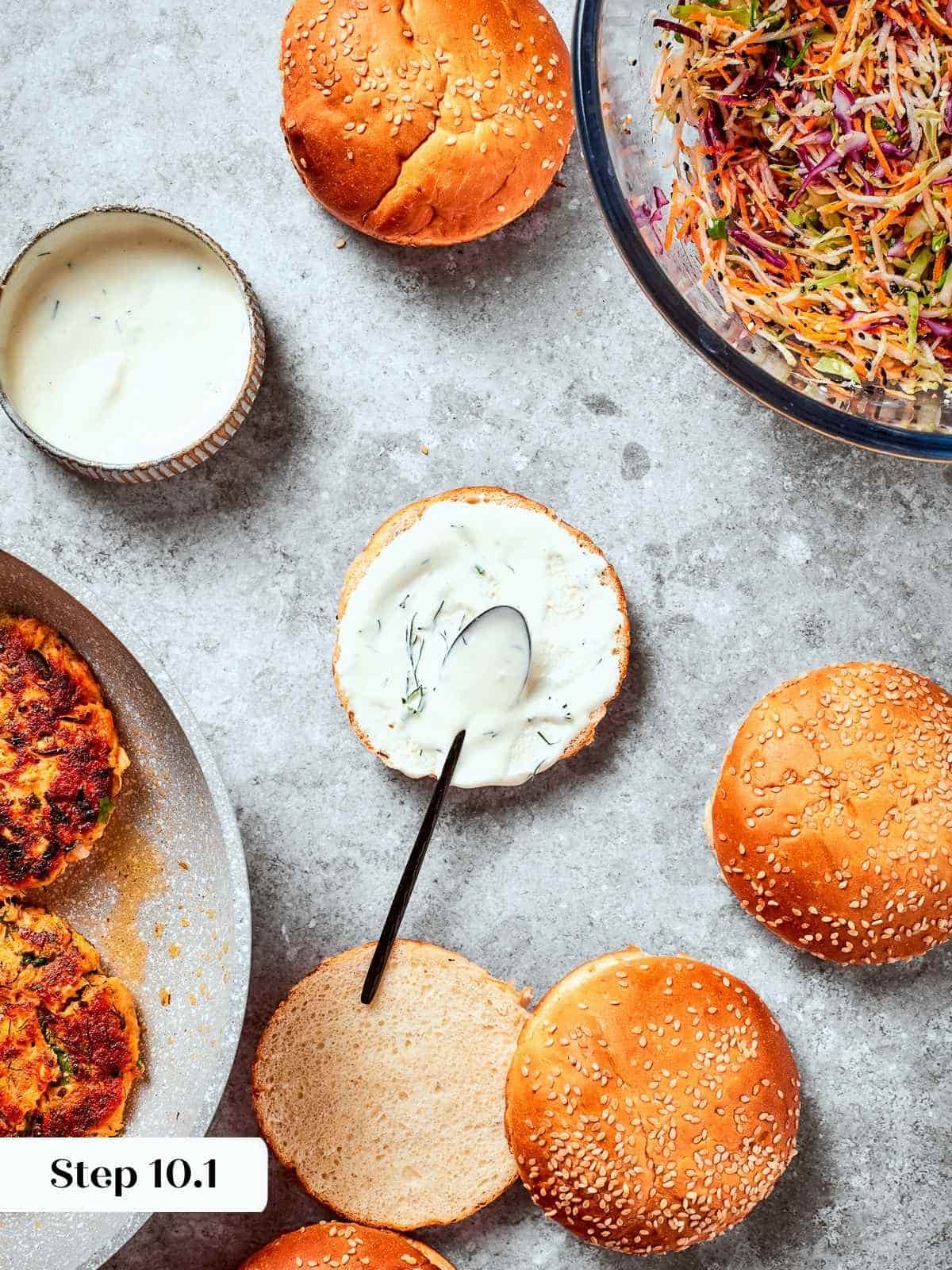 Bottom burger bun being spread with tzatziki as base for assembling a salmon burger.