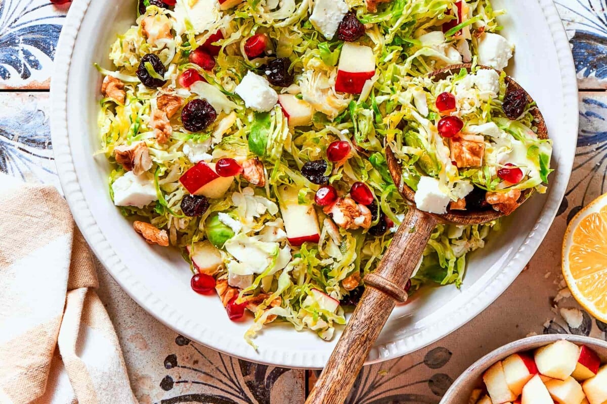 Shaved Brussels sprouts salad topped with pomegranate seeds, walnuts, and feta cheese on serving dish.