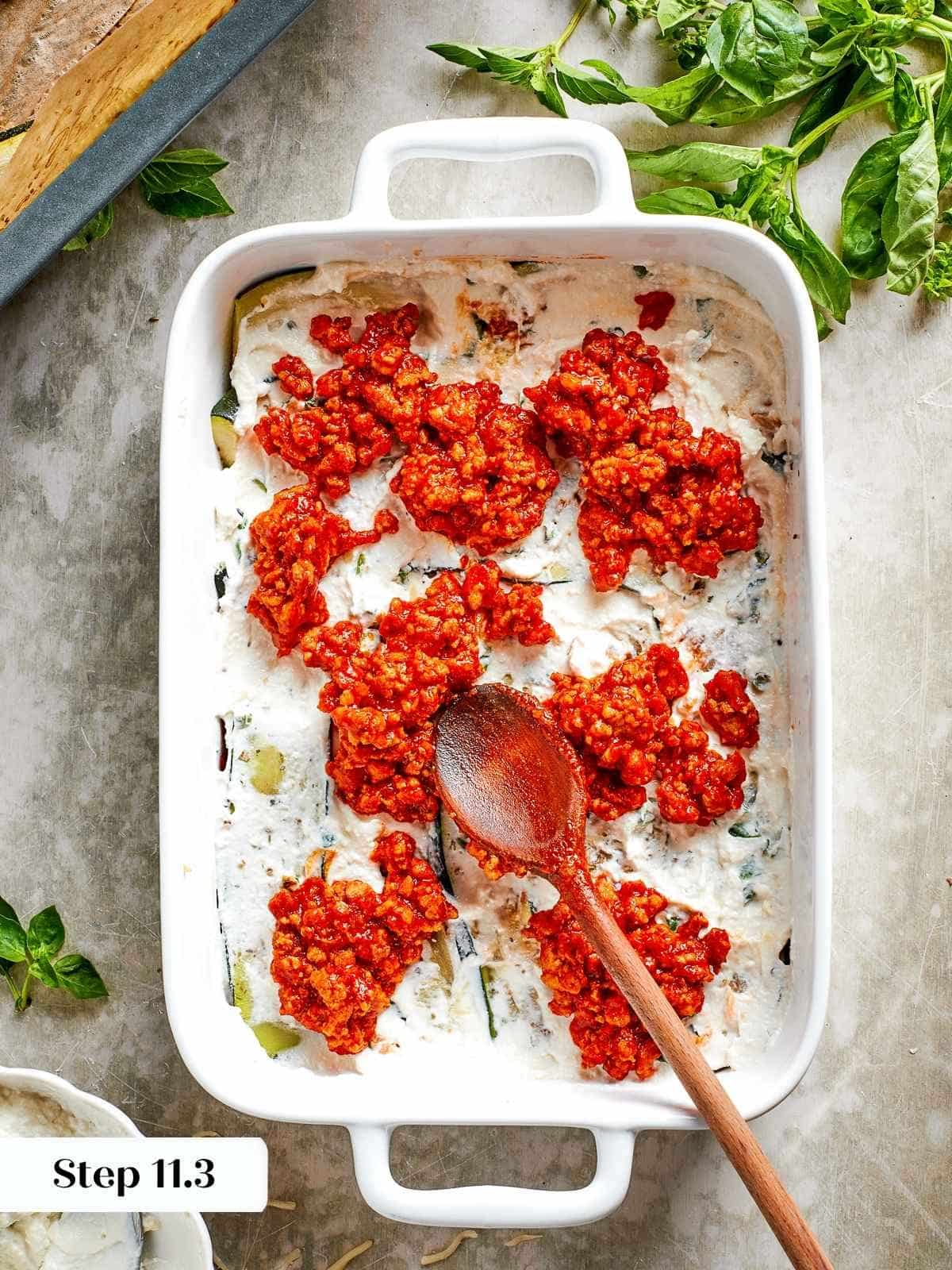 A spoon spreading tomato chicken sauce over ricotta during the zucchini lasagna layering process.