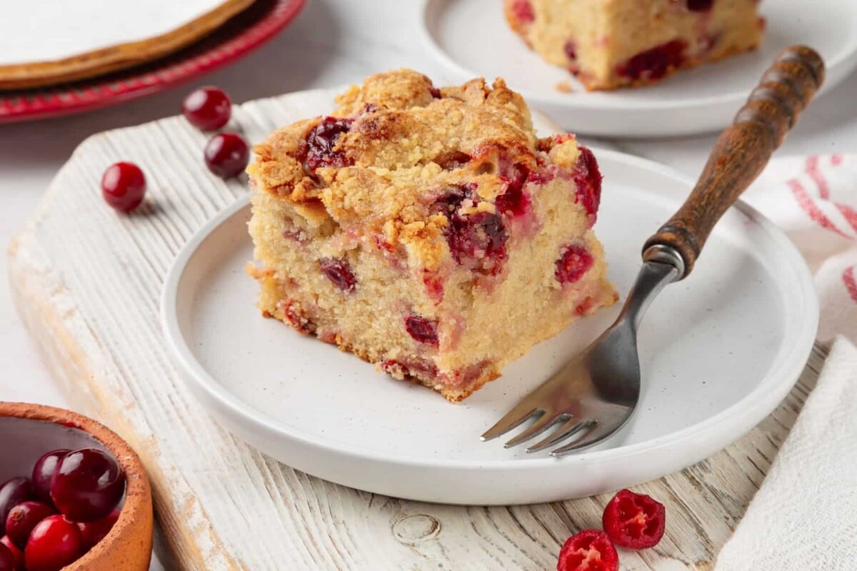 Slice of cranberry cake on a white plate showing tender crumb and baked cranberries.