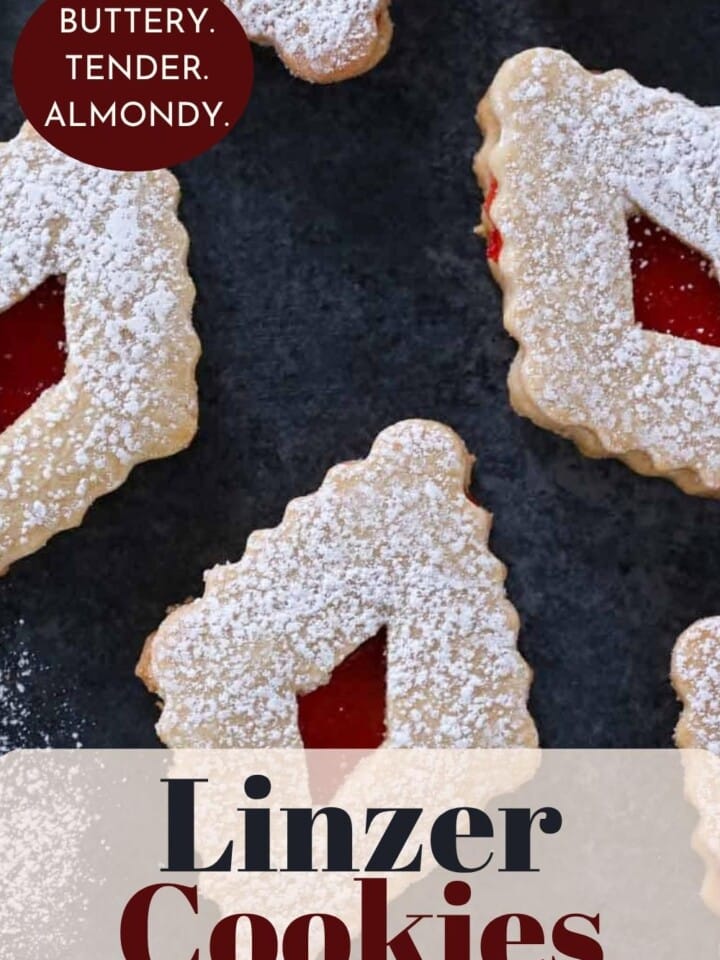 A batch of classic Linzer cookies showing golden edges and jam centers peeking through the cutouts.