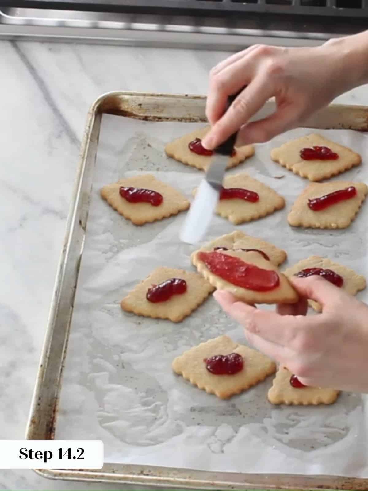 Raspberry jam being spread over baked cookie bottoms before adding the sugar-dusted tops.