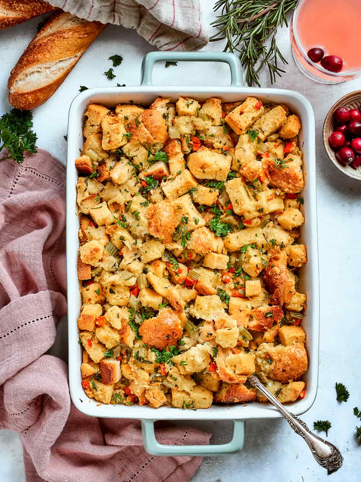 Baked cornbread dressing beside a sliced baguette and fresh rosemary on a rustic table.
