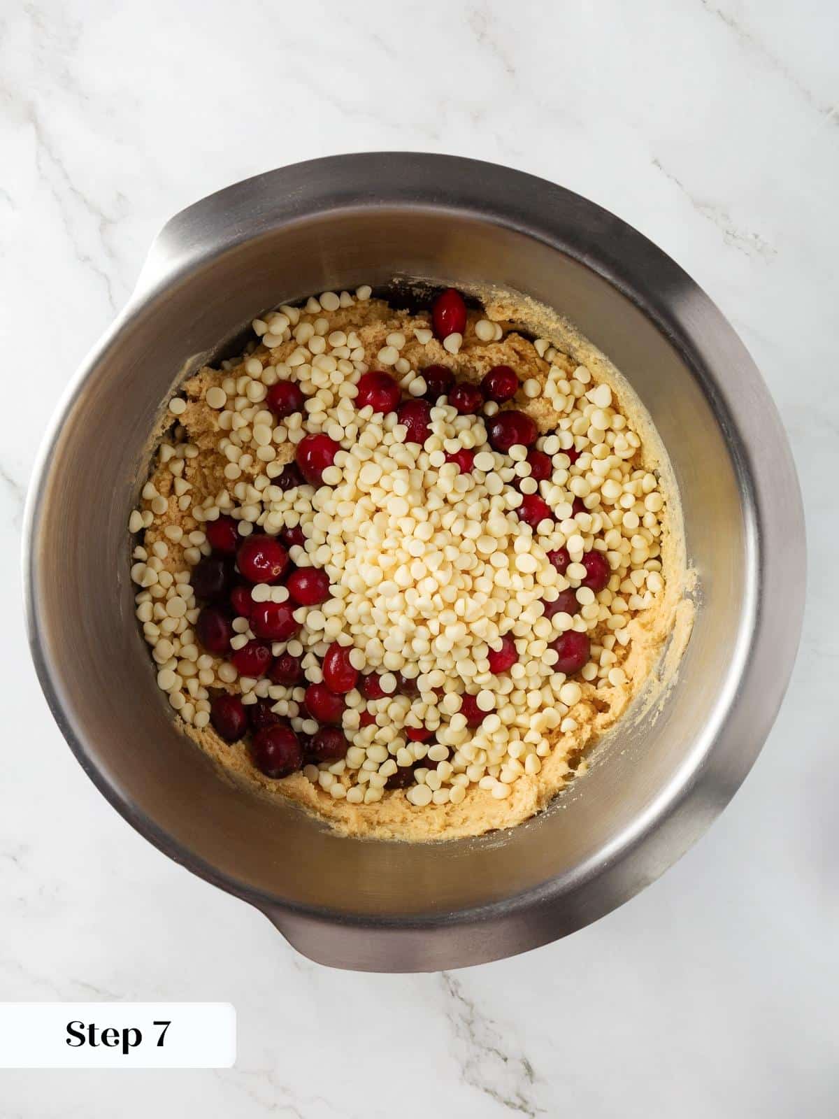 Cranberries and white chocolate chips being folded into thick cookie dough in a mixing bowl.