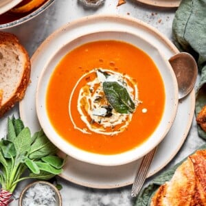 Orange pumpkin soup served with a spoon, fresh herbs, and small pieces of bread on the side.