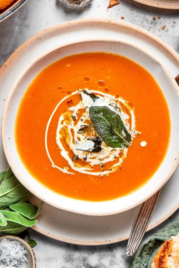 Orange pumpkin soup served with a spoon, fresh herbs, and small pieces of bread on the side.
