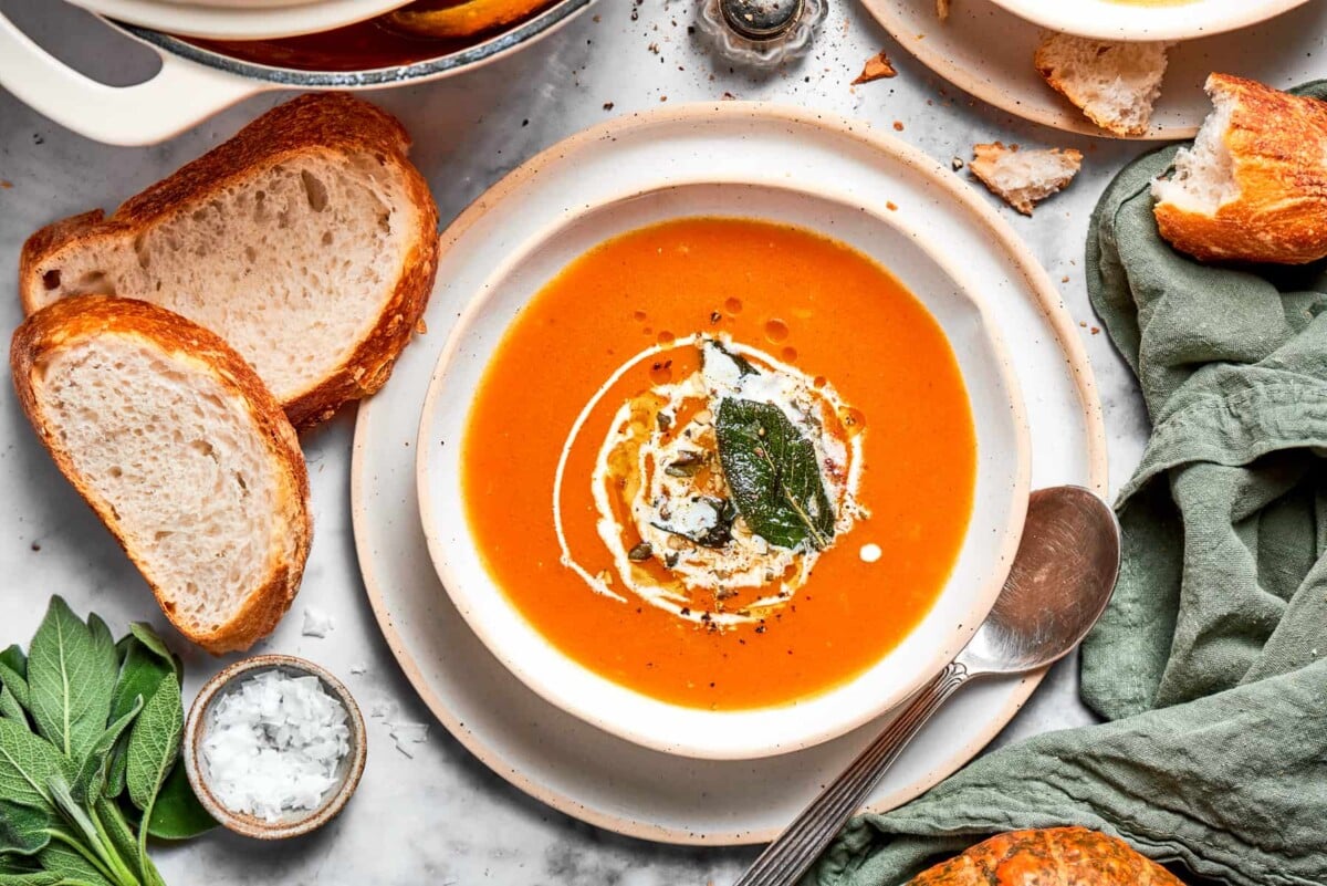 Pumpkin soup garnished lightly with herbs and served with bread and a spoon beside the bowl.