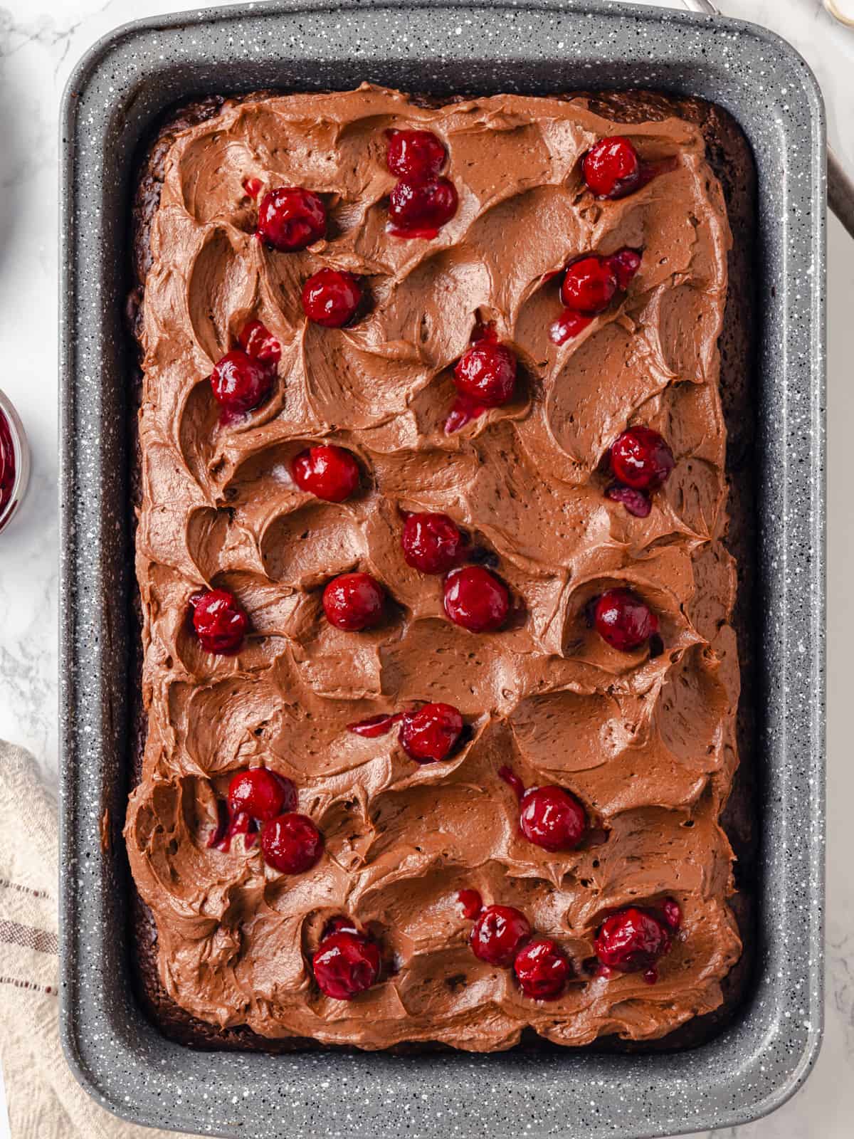 Uncut chocolate cake in rectangular dish topped with swirled frosting and cherries.