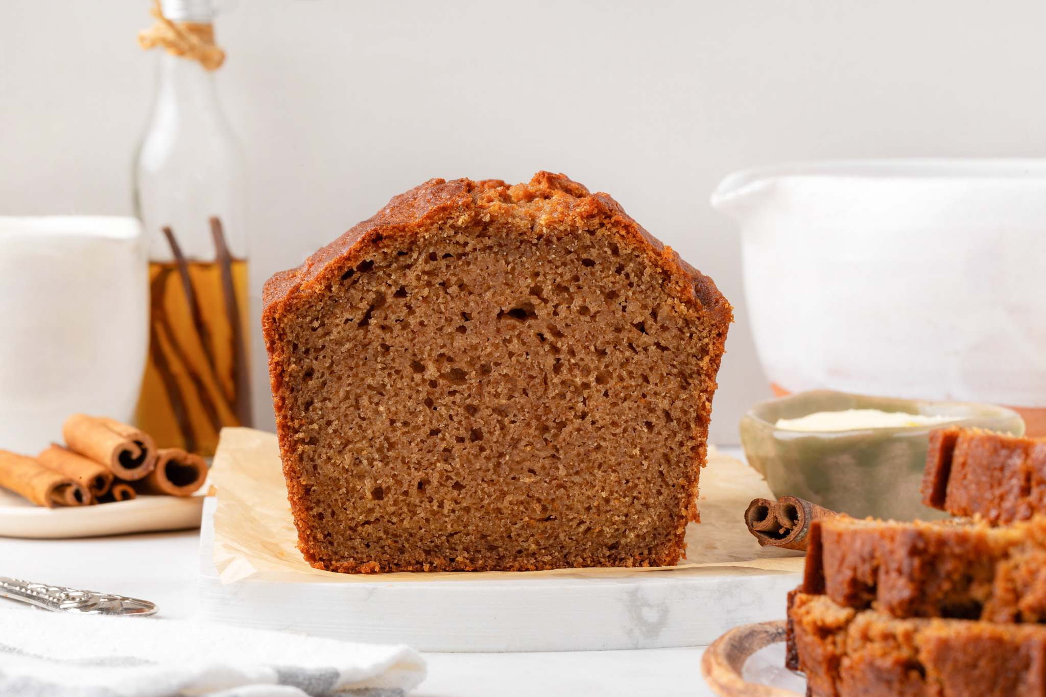 Cross-section of the loaf showing fine moist crumb framed by cinnamon sticks and a white pitcher.