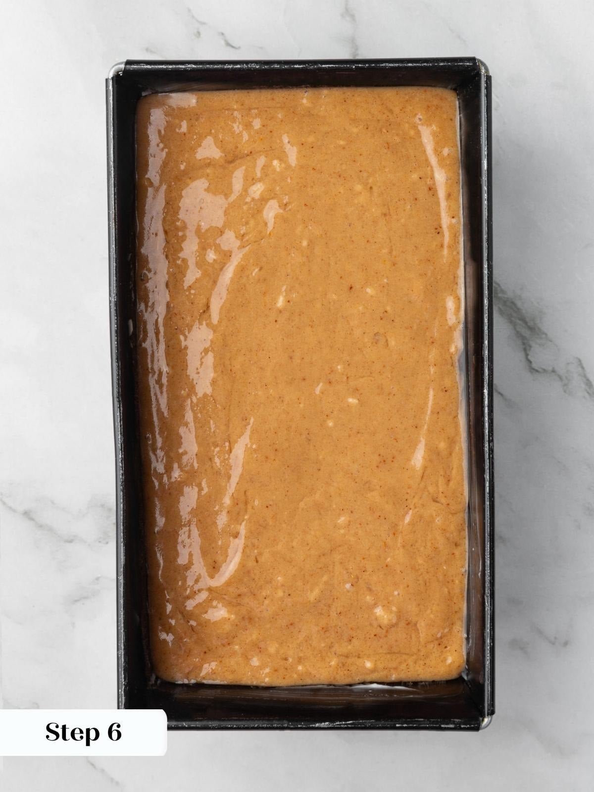 Pale orange batter spread evenly inside a dark metal loaf pan on a marble surface.
