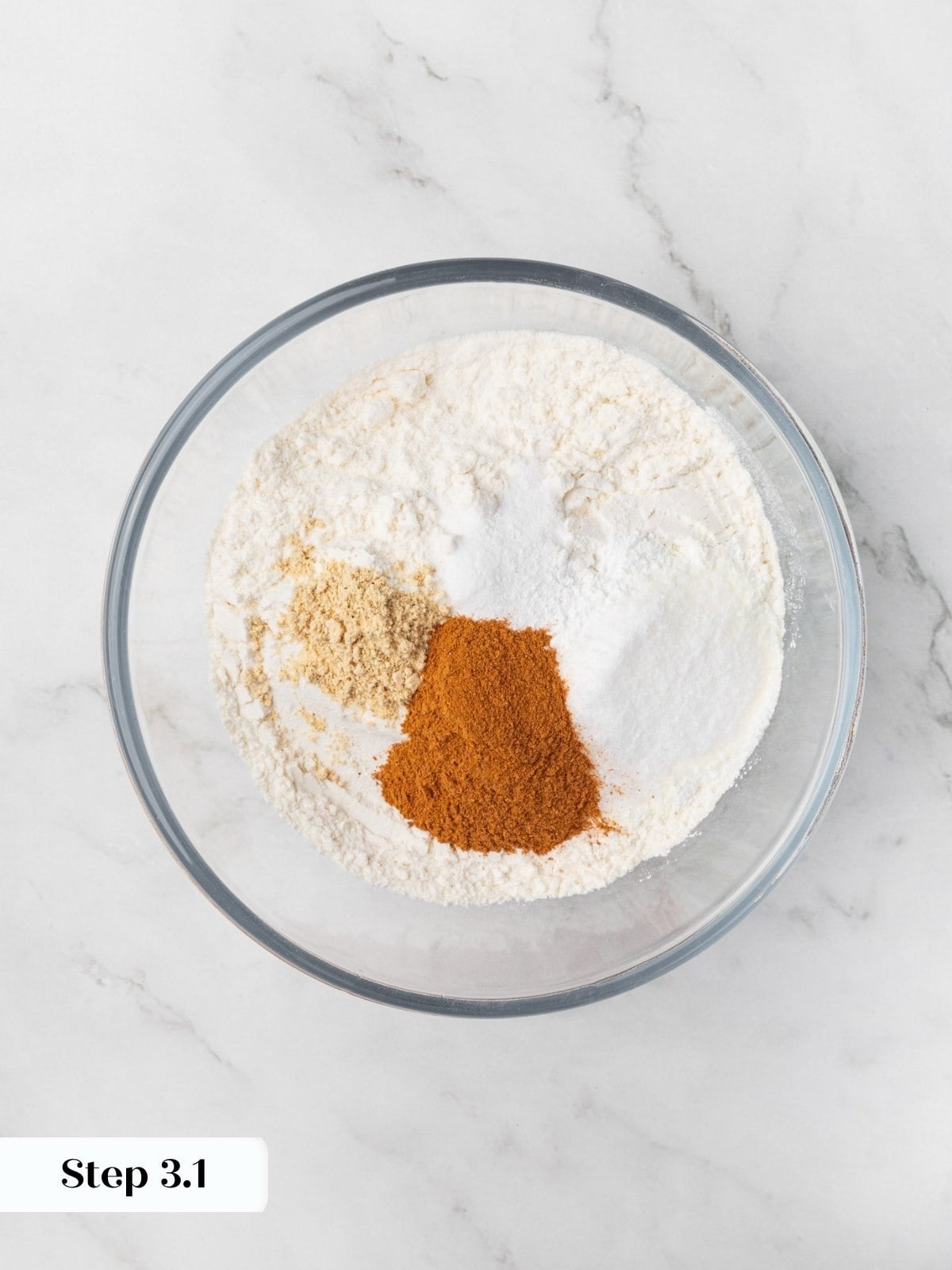 Flour with cinnamon, ginger, and white piles of leavening agents in a clear glass mixing bowl.