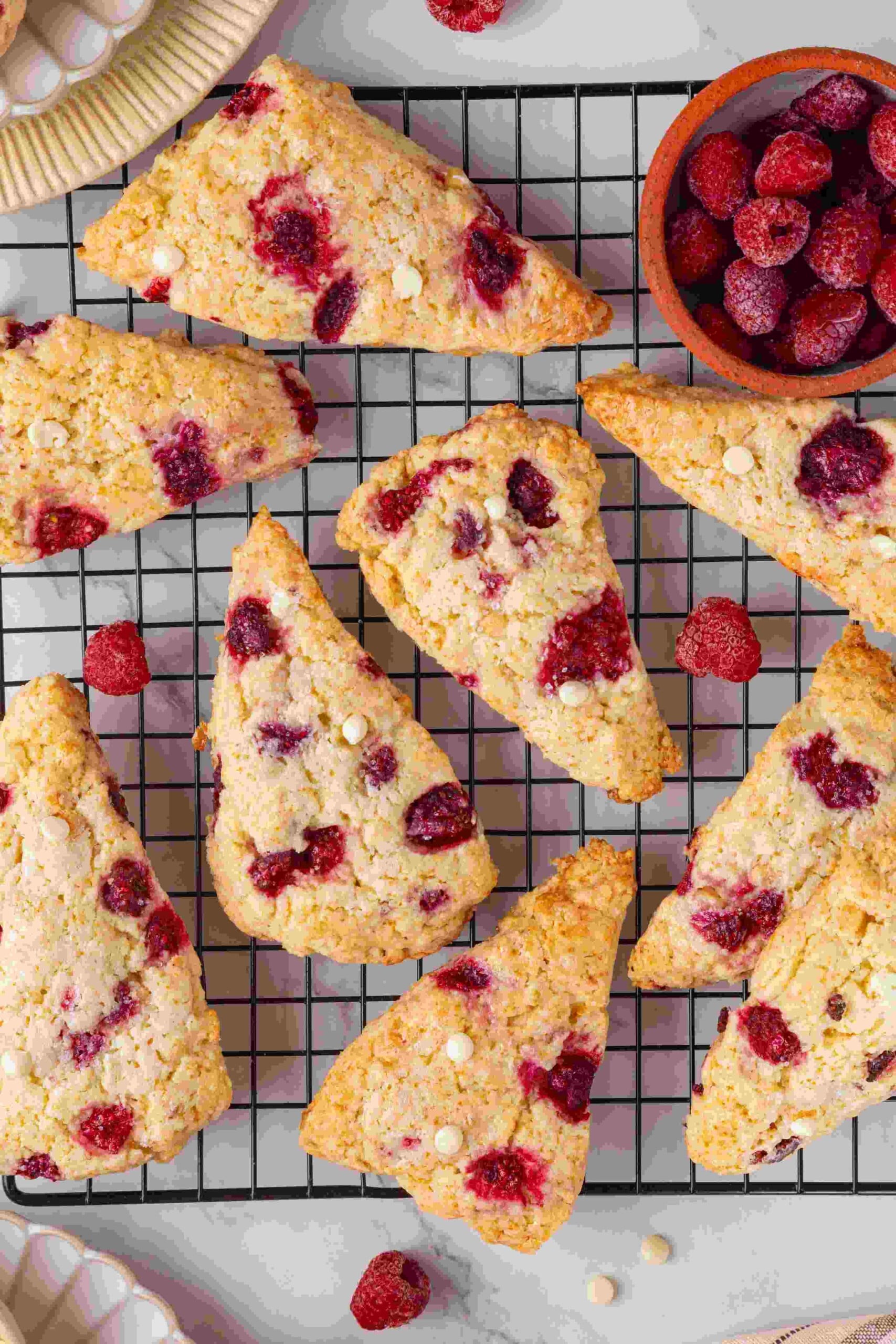 Raspberry white chocolate scones on a wire cooling rack beside a small bowl of frozen raspberries.