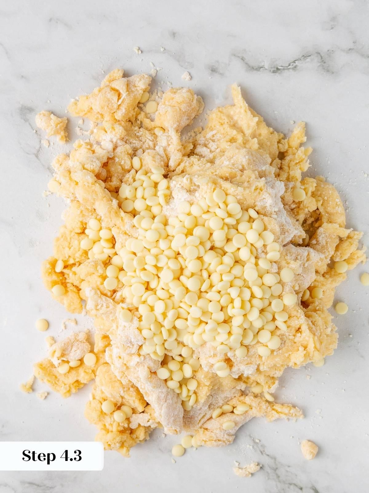 White chocolate chips piled on top of crumbly scone dough on a marble countertop before folding.