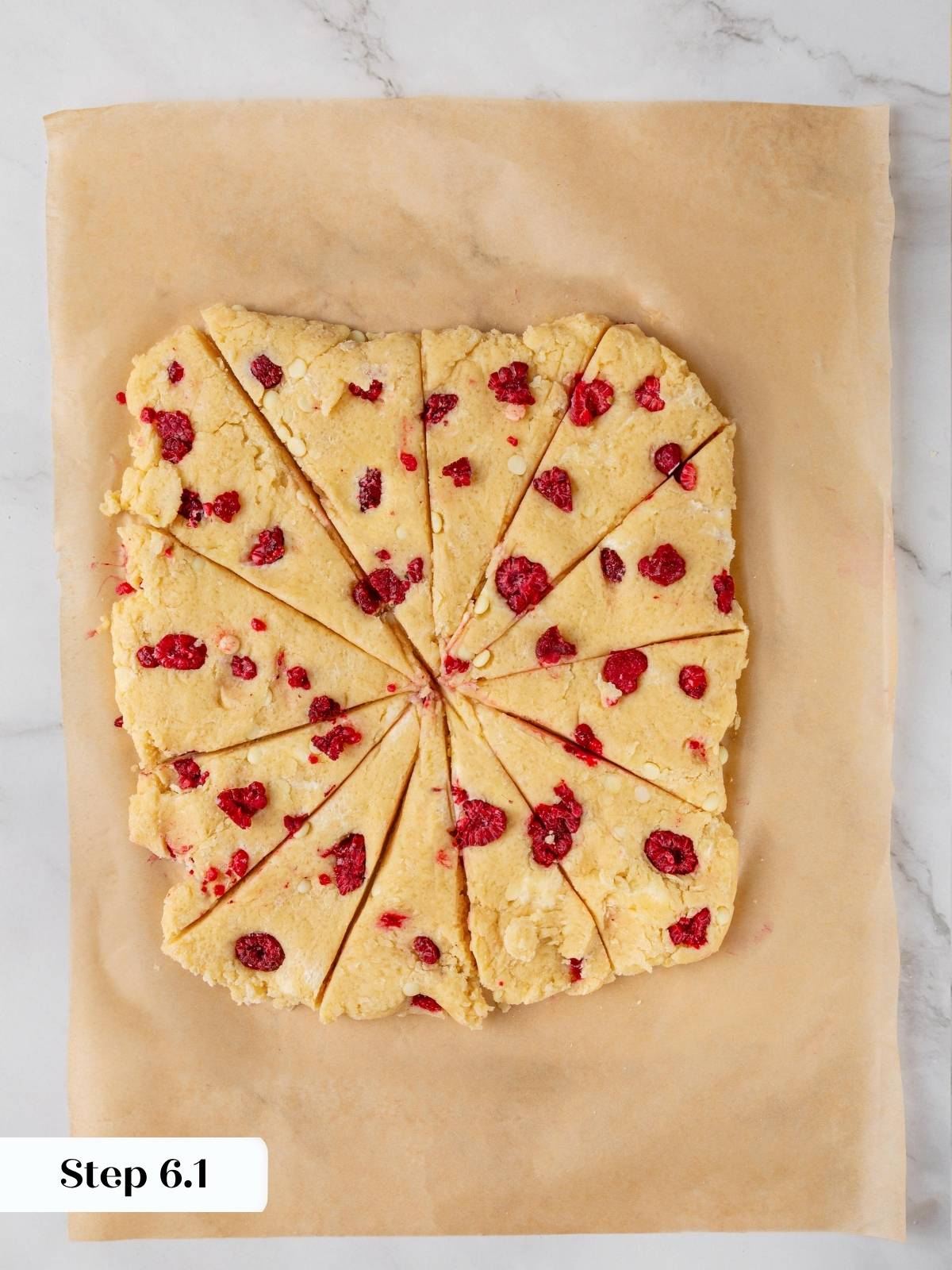 Square scone dough cut into twelve triangles on parchment paper with raspberries and white chocolate visible.