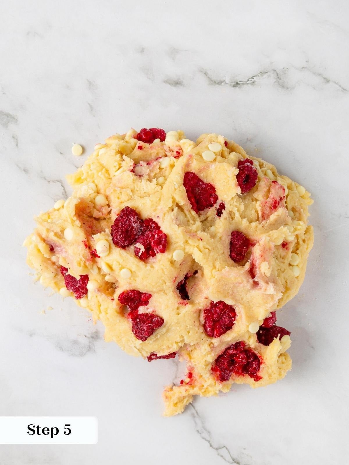 Frozen raspberries and white chocolate chips folded into pale yellow scone dough on a marble countertop.