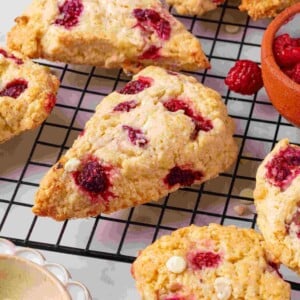 Single raspberry white chocolate scone on a wire rack with frozen raspberries in a terracotta bowl nearby.