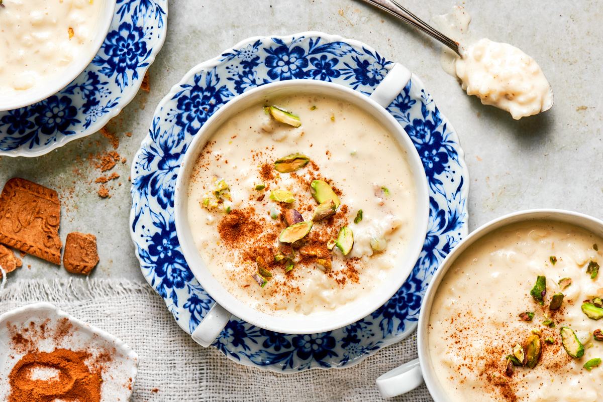 Pulled-back wide shot of rice pudding recipe with floral china, pistachios, and ground cinnamon.