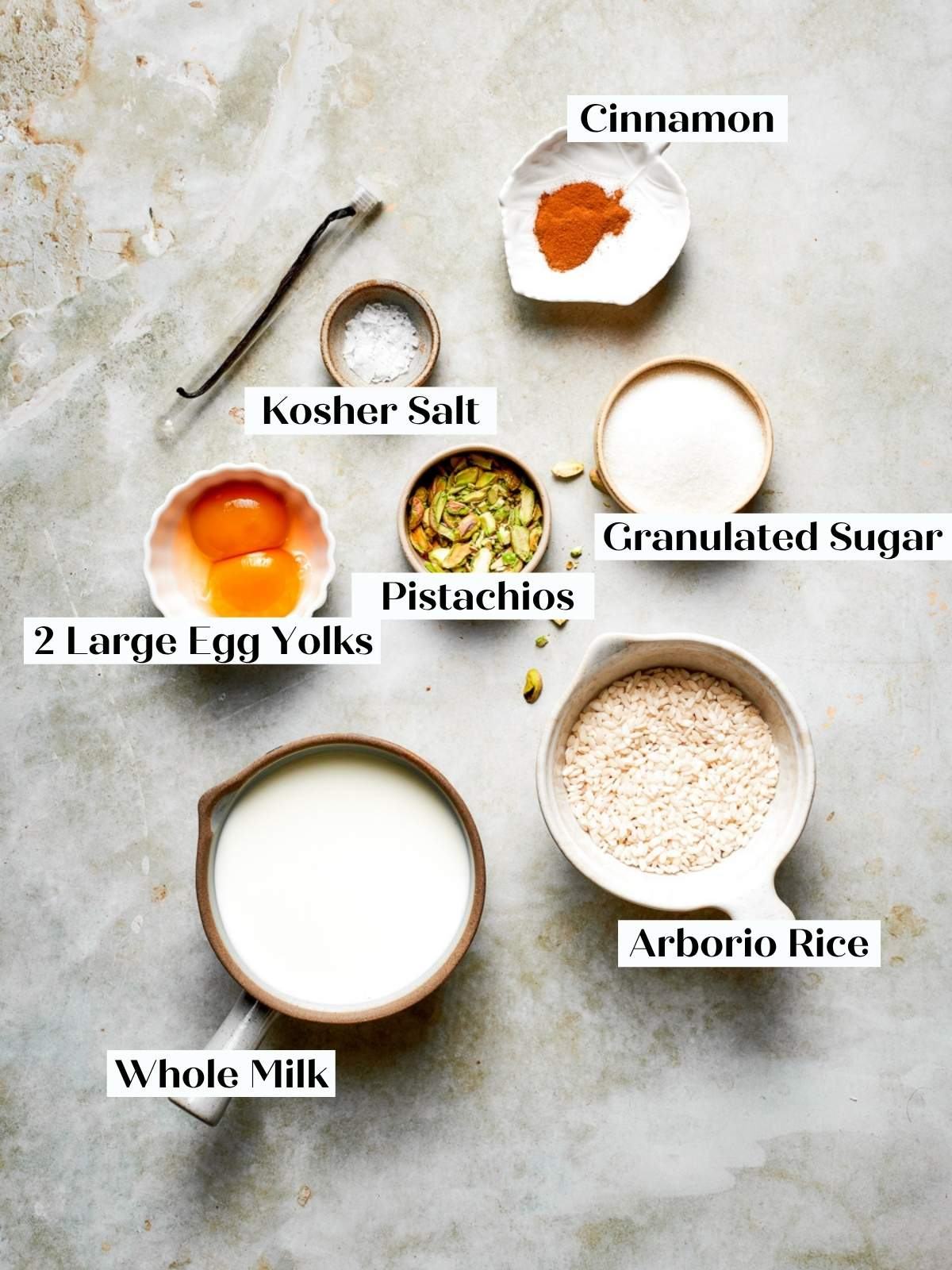 Ingredients for rice pudding recipe arranged on a grey surface including milk, arborio rice, and yolks.